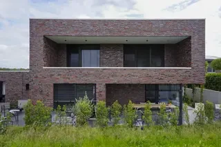 Vorderansicht eines modernen, zweigeschossigen Einfamilienhauses mit Flachdach und lebhaft gemischter rot‑braun‑grauer Klinkerfassade. Das Obergeschoss ist zurückgesetzt und bildet eine breite, schattige Loggia mit dunklen Außenjalousien vor großflächigen Fenstern. Davor liegt eine Terrasse mit Sitzbereich, eingelassen hinter einem Streifen junger, niedriger Bepflanzung.